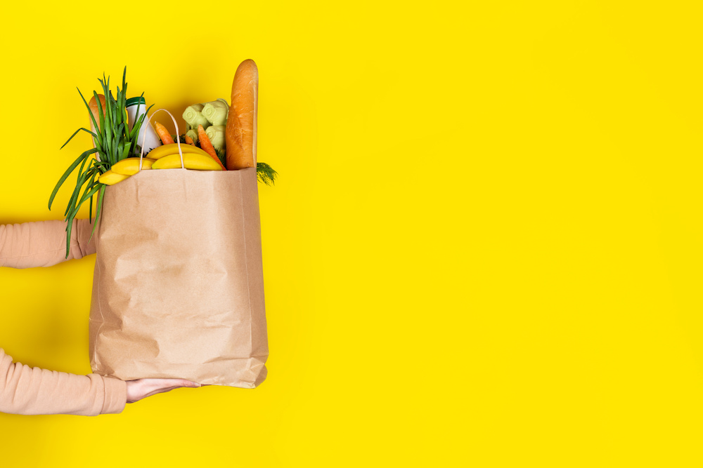 A grocery bag filled to the brim being handed over to a happy customer on top of a dazzling bright yellow background